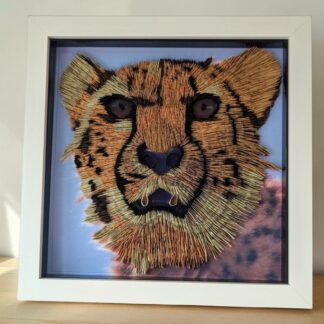 A photo of a cheetahs head looking at the camera. The photo has been quilled to make it 3D, keeping the details in the fur. It is in a white box frame.