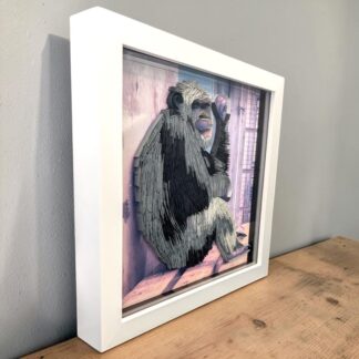 A photo of a chimpanzee sitting on a shelf. His knees are bent and he is resting his left arm on one knee and leaning his head against his hand. The photo has been quilled to make it 3D. It is seen from the left edge and is in a white box frame.