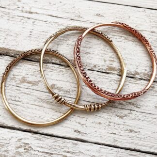 Three handcrafted copper and bronze bangles with a unique textured pattern around the outer edge. The bangles are displayed on a white background resting on a light coloured stone.