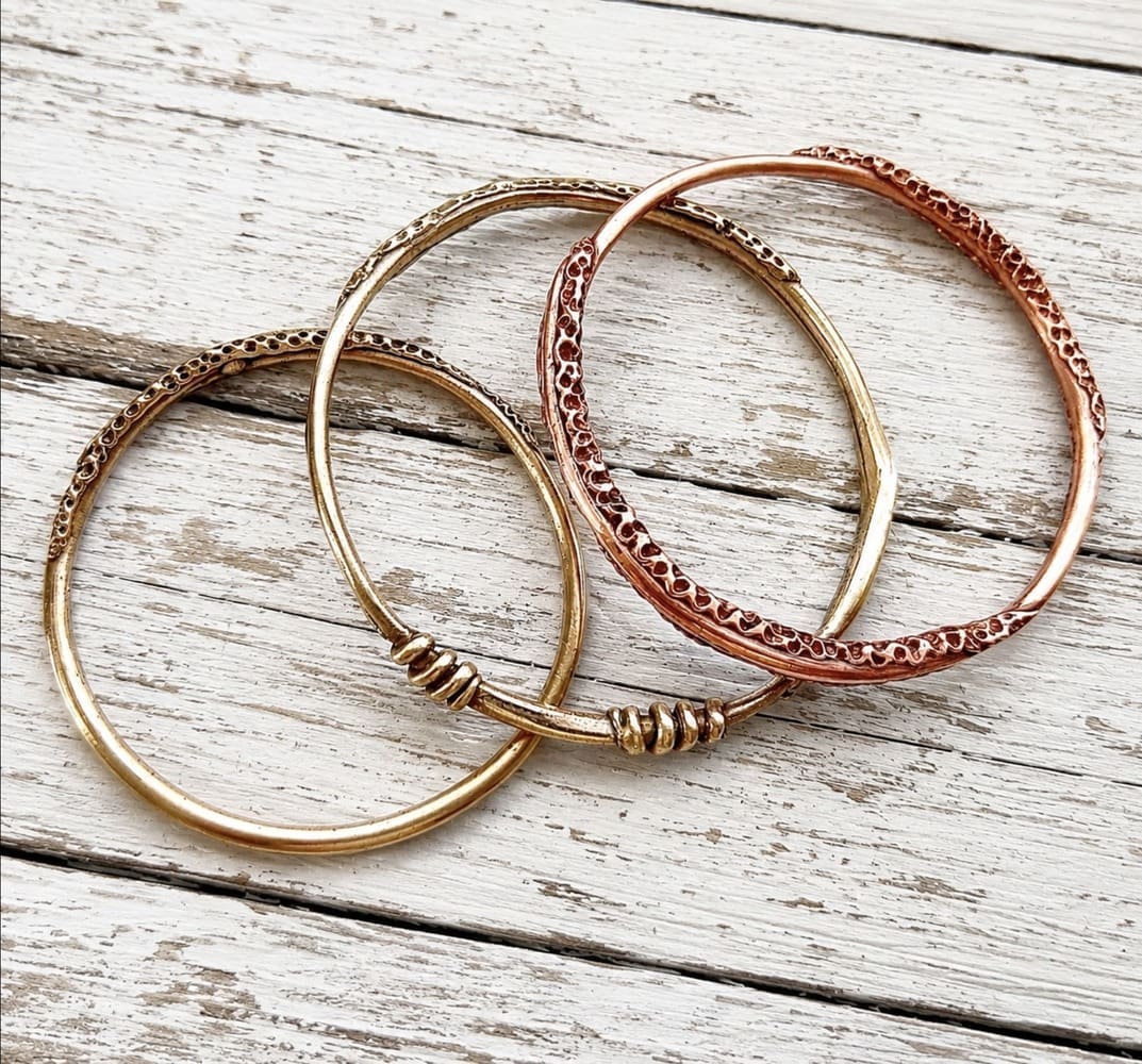 Three handcrafted copper and bronze bangles with a unique textured pattern around the outer edge. The bangles are displayed on a white background resting on a light coloured stone.