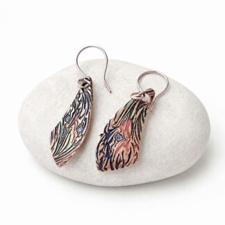 A pair of handcrafted copper drop earrings, textured and hand painted in primary colours, in a banana shape and has copper fish hook ear wires. They are displayed resting on a pale coloured stone.
