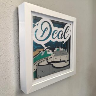 A layered picture of Deal castle in Deal Kent. It is looking from behind the castle with the beach to the left and town at the back, the background is blue mirrored card. The word Deal is written at the top of the front layer. It is in a white box frame and seen from the left side.