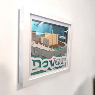 A layered paper picture of Dover Castle in Kent. The back ground is mirrored blue card and the harbour can be seen in the distance. The castle wall and main building of the castle take up most of the image. The word Dover is written at the bottom of the front layer. Looking from the left edge of the piece, it is in a white box frame.