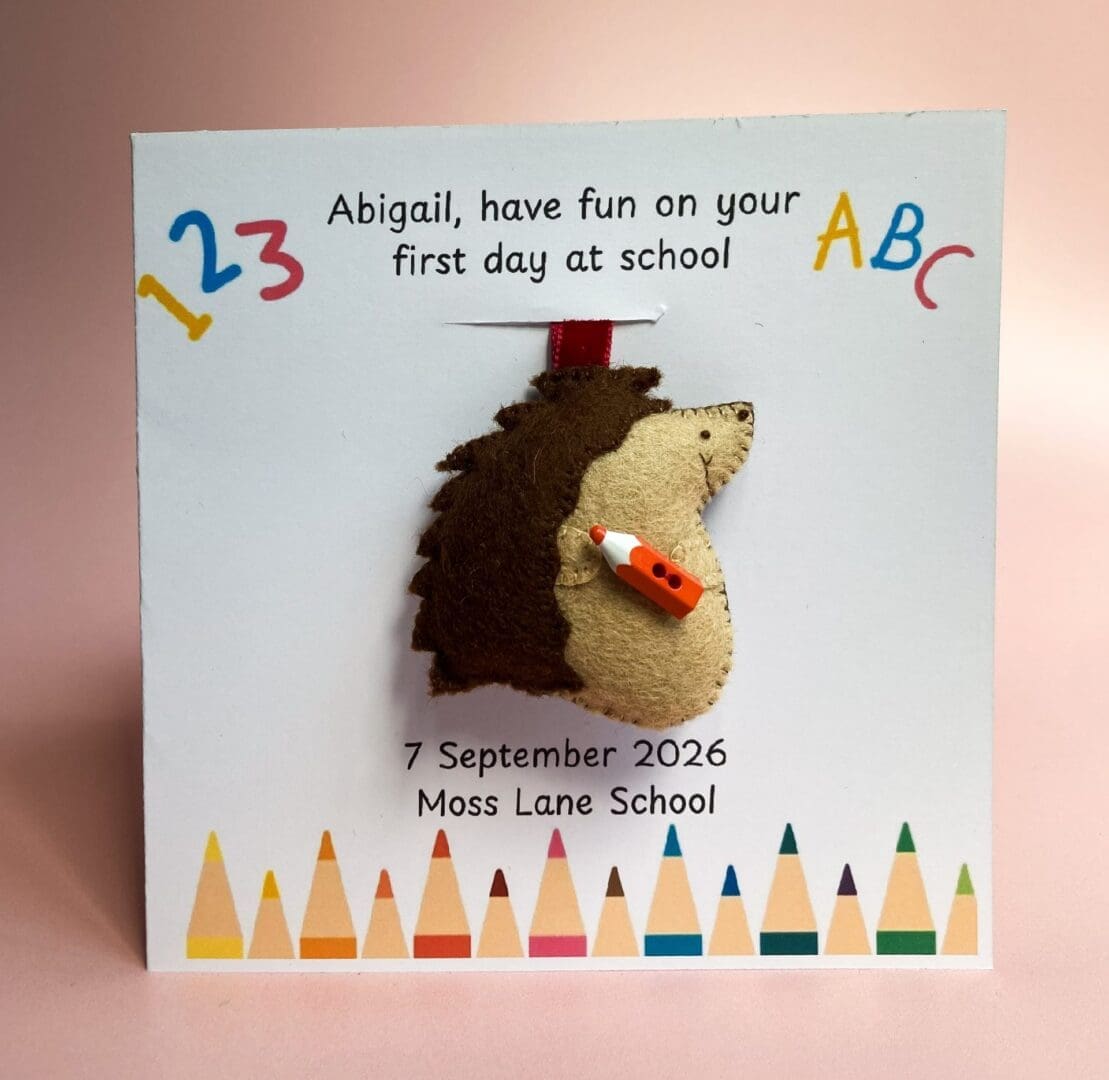 Personalised first day at school card with bright crayons at the bottom of the card, the card has a removable handmade hedgehog bag charm