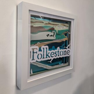 A layered paper picture of Folkestone seen from the sky, looking from the harbour arm towards the shore. The background is blue mirrored card and the front layer has Folkestone written just below the middle. The art is looked at from the left side and is in a white box frame.