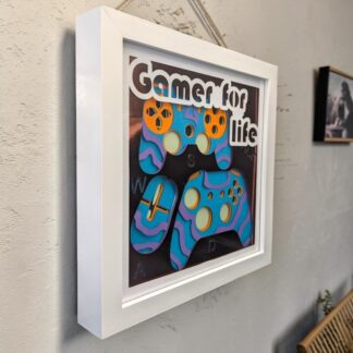 A layered paper artwork showing a computer mouse, 2 gaming controllers and the letters WASD. the front layer is mirrored card and it ahs the words 'gamer for life' written a the top. It is seen from it's left side and is in a white box frame