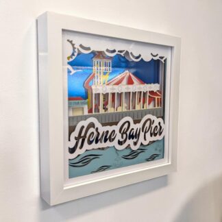 A layered artwork of the traditional carousel and helter skelter on Herne Bay Pier in Kent. The back layer is in mirrored blue card and the front layer has the words Herne Bay Pier on the bottom half. It is seen from it's left edge and is in a white box frame