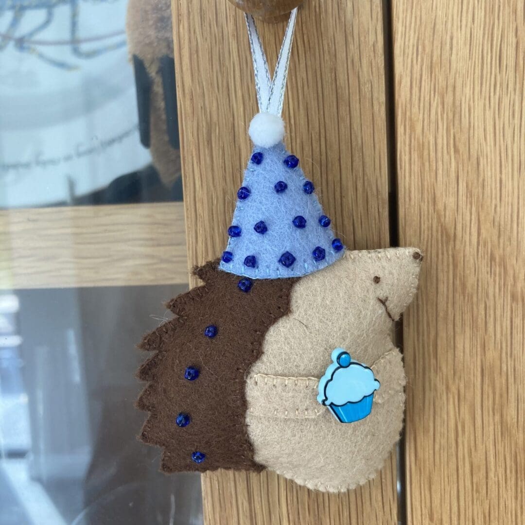 Handmade felt birthday hanging ornament with a blue party hat adorned in tiny blue beads, The hedgehog has brown pickles which have blue beads on them, The hedgehog can a cream tummy and hands. The hedgehog is holding a blue cupcake ready for a birthday party.