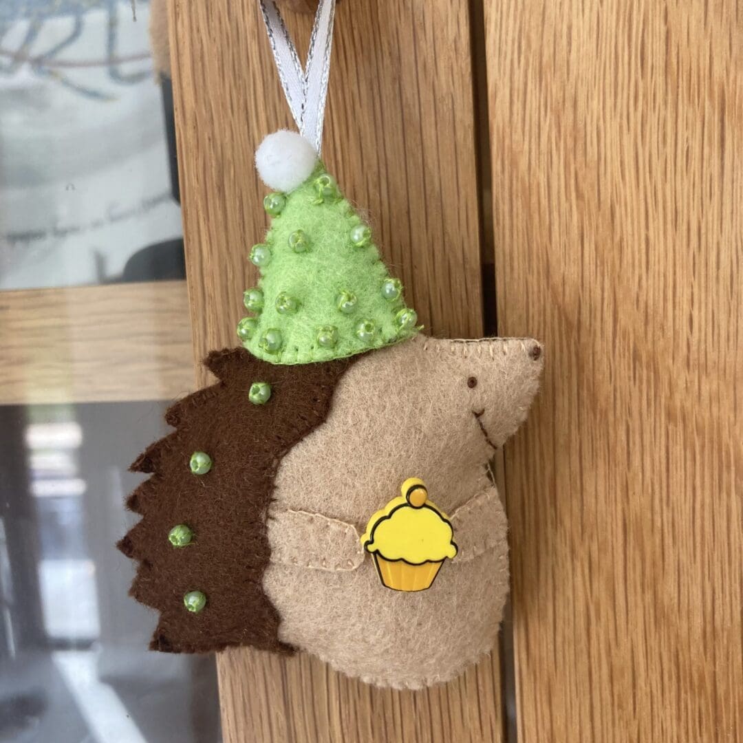 Handmade felt birthday handing hedgehog ornament. This hedgehog has brown prickles, a cream tummy and is wearing a green party hat. The hedgehog is holding a yellow birthday cupcake ready for a party. The hedgehog can be hung up and kept as a special birthday momento.
