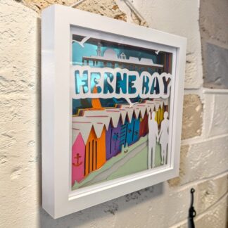 A layered paper artwork of Herne Bay in Kent. Looking from the hills over colourful the beach huts toward the Pier in the distance. The front layer has two figures walking hand in hand one is pointing towards the Pier, the words Herne Bay is written near the top of the image. The picture is seen from the left side and is in a white box frame.