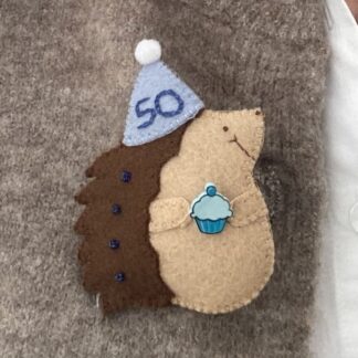 Handmade felt hedgehog birthday badge, the hedgehog is wearing a blue party hat which has 50 hand sewn on to it in dark blue. The hedgehog is carrying a blue cupcake and has blue beads sewn on to it's brown prickles