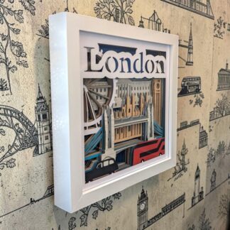 A London themed layered paper art, showing the London eye, Buckingham palace, Tower Bridge, House's of Parliament, a London bus and taxi. The picture has a blue mirror backing and the front layer says 'London' across the top. It is seen from it's left edge and is in a white box frame