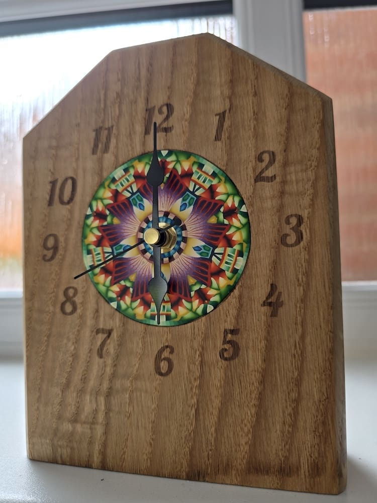 Handcrafted solid oak mantel clock with laser-engraved numerals and colourful millefiori-inspired polymer clay centrepiece artwork by Bridget Derc, featuring black hands and sharp chamfered edges.