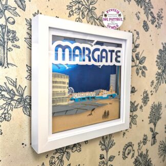 A layered paper artwork on Margate in Kent. It has a mirrored blue backing and shows the seafront buildings and steps to the beach. The front layer says 'Margate' at the top. The image is seen from it's left side and is in a white box frame.