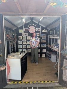 A photo of myself stood in the middle of my shop on Herne Bay Pier, There is a large circular sign above me saying 'Pretty  not purrfect crafts. There is a large display of framed artworks behind me, a desk in front of me to the left.  Hanging decorations on a rack on the left wall and shelves of coasters on the right wall.