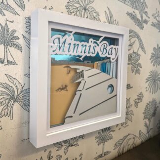 A layered paper artwork of Minnis Bay beach in Kent. It shows the promenade and blue coloured beach huts, in front of a mirrored blue background. The front layer has Minnis Bay written at the top. It is seen from it's left edge and is in white box frame