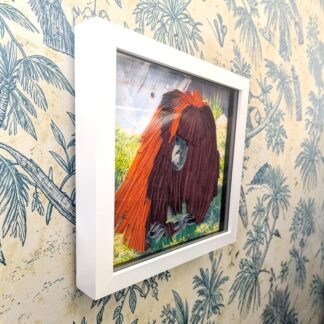 A quilled photo of an Orangutan walking on grass. It is seen from it's left edge and is in a white box frame