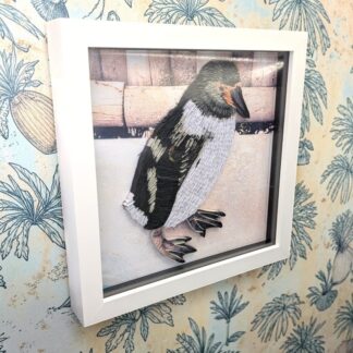 A quilled photo of a penguin stood by a fence. It is seen from the left edge and in a white box frame