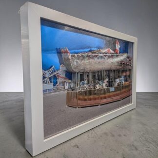 A 3D decoupage image of the carousel and helter skelter on Herne Bay Pier in Kent. It is made from multiple cut photos, layered on top of each other. This is seen from the left side of the piece and is in a white box frame.