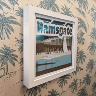 A layered paper artwork of Ramsgate in Kent. Looking across the harbour towards the arches and the businesses in the cliffside. It is seen from it's left edge and in a white box frame.
