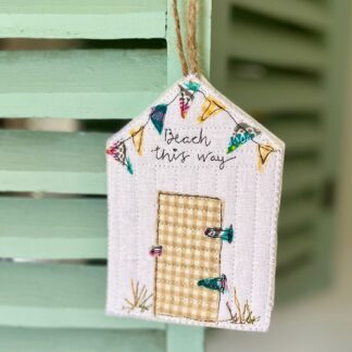 A hanging decoration of a beach hut with appliqued detailand pretty colourful bunting. Hangs on blue louvre doors. Reads beach this way.