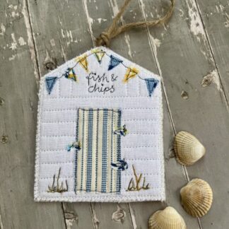 A beach hut shaped hanging decoration laying on a bench. Features blue and yellow bunting and a striped door with brown string hang to hang.