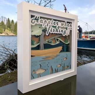 A layered paper artwork of a person stood in a small wooden boat on a river. They are fishing, below them are fish in the water and the background has woodland and mirrored blue layer. The top layer has the words ' happier when fishing' written at the top with a Kingfisher perched next to them. This picture is seen from it's left side and is in a white box frame.