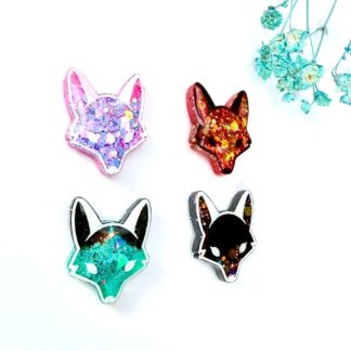 Four handmade fox and wolf glitter resin pin badges in pink holographic, classic fox, teal galaxy and shadow variations by Copper Magpie Designs Liverpool. Displayed on a white background with blue flowers. A perfect handmade gift.