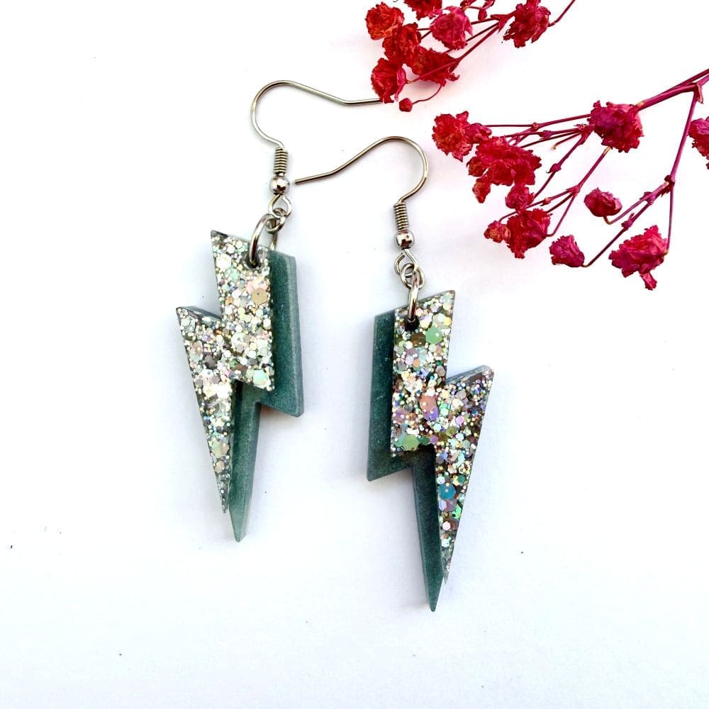 Flat lay of holographic glitter lightning bolt earrings, bold statement jewellery handmade in Liverpool, on a white background with pink flowers.