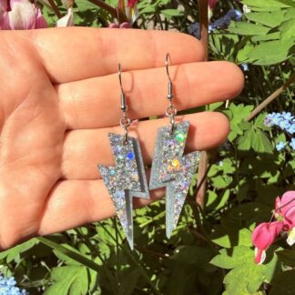 Holographic glitter lightning bolt resin earrings handmade in Liverpool by Copper Magpie Designs UK, displayed in full sun on a hand for scale with flowers in the background.