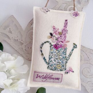 Pink and purple liberty floral fabric watering can applique hanging on wool cream felt featuring two additional applique flowers and free motion stitched text of wildflowers. Hung with a rustic twine. Flower and garden lover small gift.