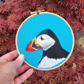 Hand embroidered puffin on a bright blue fabric background, framed in a 6 inch bamboo embroidery hoop.