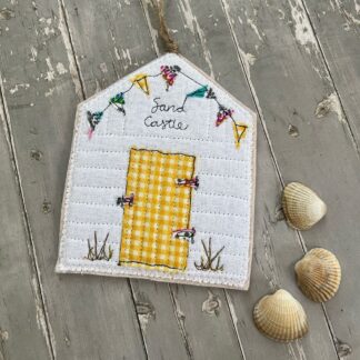A beach hut shaped felt hanging decoration with the words sand castle embroidered on the front. Has a yellow gingham door and colourful bunting appliqued.